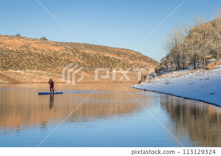 male paddler in red drysuit is paddling a stand up paddleboard on mountain lake in Colorado, winter scenery 113129324