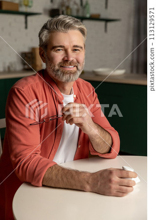 Middle aged bearded man holding glasses sitting at table at home 113129571