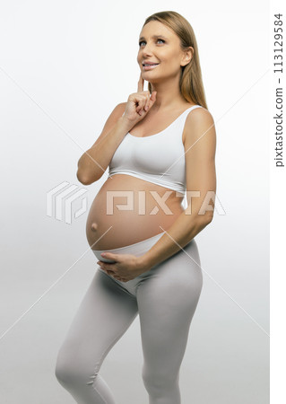 Blonde pregnant woman in white clothes looking thoughtful 113129584