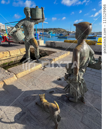 A wonderful monument on the shore of Marsaxlokk, Malta: a young son and daughter meet a father who is carrying two wicker baskets full of caught fish. 113129966