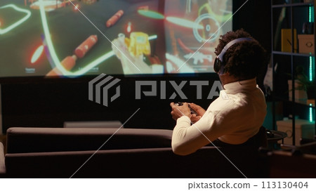 African american man watching Video on demand channel on widescreen television set in neon lit living room. Cord cutter entertained by streaming service program broadcasted on smart TV in home theatre African american man watching Video on demand channel on widescreen television set in neon lit living room. Cord cutter entertained by streaming service program broadcasted on smart TV in home theatre 113130404