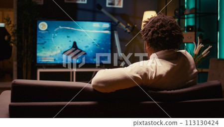 BIPOC gamer plays game with spaceship shooting laser streams using crosshair overlay to hit enemies. Ray tracing graphics and high FPS scifi videogame displayed on television set, zoom in shot BIPOC gamer plays game with spaceship shooting laser streams using crosshair overlay to hit enemies. Ray tracing graphics and high FPS scifi videogame displayed on television set, zoom in shot 113130424