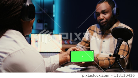 Man invited to podcast watching internet clips on green screen smartphone with host in studio, discussing content. BIPOC guest reacting to videos on mockup mobile phone during online comedy show Man invited to podcast watching internet clips on green screen smartphone with host in studio, discussing content. BIPOC guest reacting to videos on mockup mobile phone during online comedy show 113130436