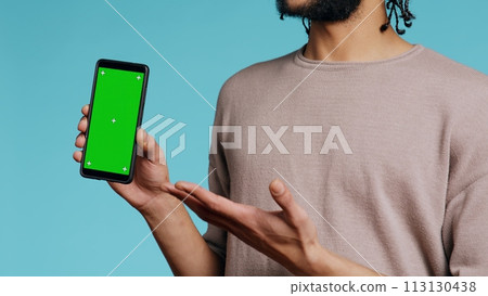Happy man pointing towards green screen mobile phone, showing thumbs up sign. Cheerful person presenting chroma key cellphone, doing gesturing, isolated over studio background, camera B close up 113130438