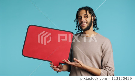 Smiling BIPOC man holding red speech bubble sign of empty copy space for message. Joyous person presenting thought bubble cardboard used as promotion concept, isolated over studio background 113130439