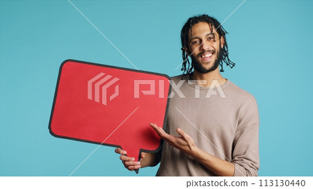 Happy man holding red speech bubble sign of empty space for text. Cheerful Middle Eastern person presenting thought bubble cardboard, isolated over blue studio background 113130440