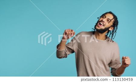Happy Middle Eastern dancer having fun, dancing on rhythm, enjoying leisure time. Cheerful BIPOC man entertaining himself doing funky movements on music, studio background, camera A 113130459