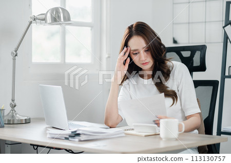 Concerned young businesswoman carefully examines paperwork at her office desk, showing signs of stress. 113130757