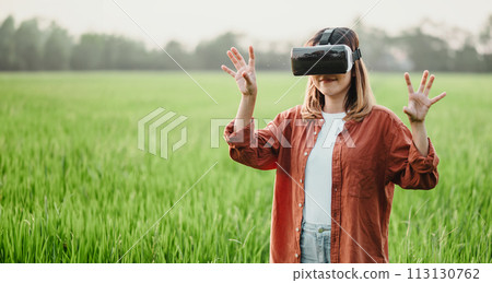 Woman gestures with her hands while engaged with a virtual reality headset amid a vibrant green field. 113130762