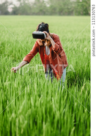 Woman in a rustic orange jacket reaches out while wearing a VR headset, surrounded by tall, lush green grass. 113130763
