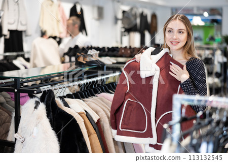 Girl stopped near showcase with outerwear, holds fur coat hanger in hands and examines product Girl stopped near showcase with outerwear, holds fur coat hanger in hands and examines product 113132545