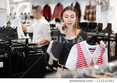 While shopping, girl looks at blouse and chats on phone with friend, discusses latest news. 113132561