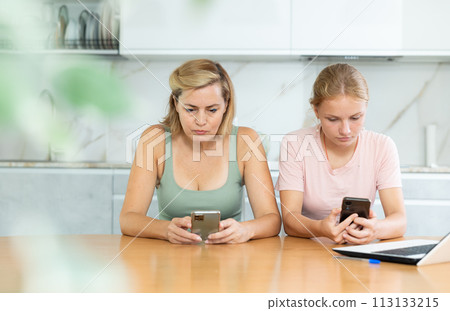 Woman and teenage daughter sitting in kitchen with phones in their hands and intently reading news Woman and teenage daughter sitting in kitchen with phones in their hands and intently reading news 113133215