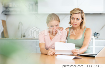 Upset teenage girl reding papers with worried mother in home kitchen 113133313