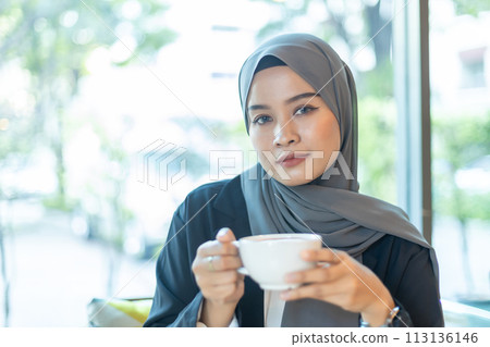 beautiful muslim woman wear hijab holding coffee cup looking at camera sitting at cafe. beautiful muslim woman wear hijab holding coffee cup looking at camera sitting at cafe. 113136146