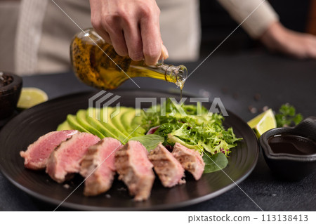Woman pours cooking olive oil on a salad on a plate with grilled cooked piece of tuna fillet Woman pours cooking olive oil on a salad on a plate with grilled cooked piece of tuna fillet 113138413