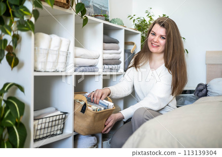 Modern housewife putting neatly folded towels, duvet covers into wicker natural eco-friendly basket Modern housewife putting neatly folded towels, duvet covers into wicker natural eco-friendly basket 113139304