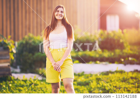 Yellow Bermuda shorts are worn by Caucasian young brunette woman who poses for photograph smiling while standing in backyard of country house at end of day. 113142188