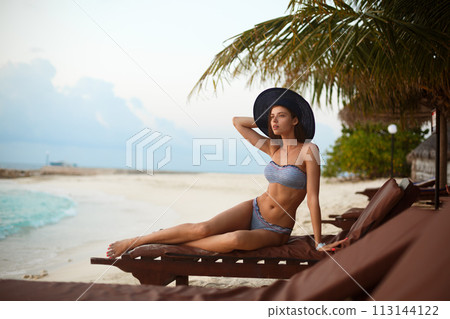 Young woman relaxing in a deck chair on a tropical beach at sunrise or sunset with stylish straw hat on, girl on a beach sun bed chilling near ocean and palms on Maldives Young woman relaxing in a deck chair on a tropical beach at sunrise or sunset with stylish straw hat on, girl on a beach sun bed chilling near ocean and palms on Maldives 113144122