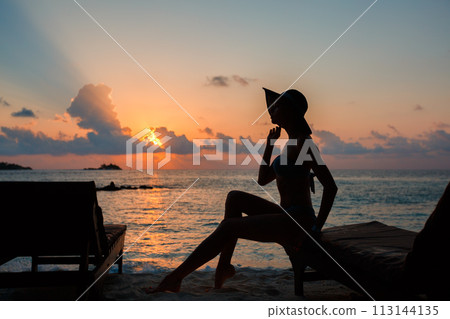 Silhouette of a beautiful slender girl on a sunset background and sun loungers on the ocean shore. Woman watch skyline on sunrise on near deck chair at the sea beach. Vacation Concept. Silhouette of a beautiful slender girl on a sunset background and sun loungers on the ocean shore. Woman watch skyline on sunrise on near deck chair at the sea beach. Vacation Concept. 113144135