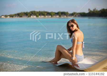 Young woman relaxing in a modern deck chair on a tropical beach with glasses on. Girl is sitting on a beach sun bed chilling near ocean, palms and bungalows. Copy space 113144192