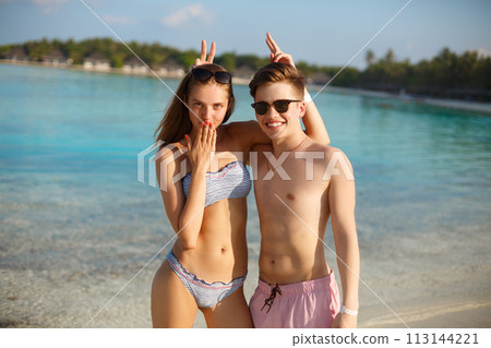 Happy young couple have fun and relax on the beach. Man and woman posing for photo and fool around. Bungalows of spa resort, palms, and blue sea on background. Family travel concept Happy young couple have fun and relax on the beach. Man and woman posing for photo and fool around. Bungalows of spa resort, palms, and blue sea on background. Family travel concept 113144221
