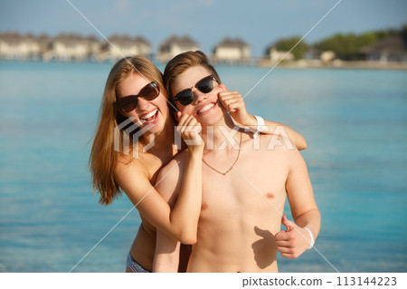 Happy young couple have fun and relax on the beach. Woman makes man smile holding cheeks with hands. Bungalows of spa resort, palms, and blue sea on background. Family travel concept 113144223
