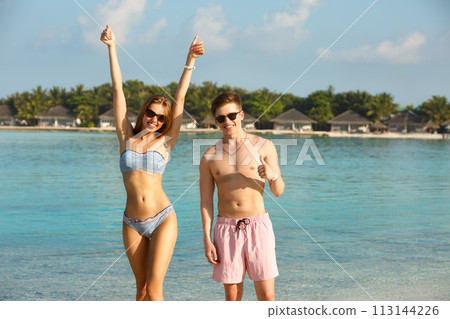 Happy young couple have fun and relax on the beach. Man and woman show thumbs up enjoying vacation near ocean. Bungalows of spa resort, palms, and blue lagoon on background. Family travel concept Happy young couple have fun and relax on the beach. Man and woman show thumbs up enjoying vacation near ocean. Bungalows of spa resort, palms, and blue lagoon on background. Family travel concept 113144226
