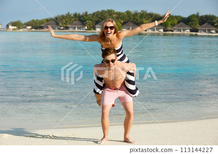 Happy young lovely couple have fun and relax on the beach. Man holds woman showing plane on his back. Bungalows of spa resort, palms, and blue lagoon on backgrpund. Family travel concept Happy young lovely couple have fun and relax on the beach. Man holds woman showing plane on his back. Bungalows of spa resort, palms, and blue lagoon on backgrpund. Family travel concept 113144227