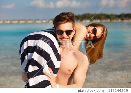 Happy young lovely couple have fun and relax on the beach. Man holds woman on his shoulder. Bungalows of spa resort, palms, and blue lagoon on backgrpund. Family travel concept Happy young lovely couple have fun and relax on the beach. Man holds woman on his shoulder. Bungalows of spa resort, palms, and blue lagoon on backgrpund. Family travel concept 113144229