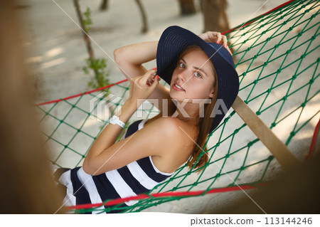 Woman relaxing in the hammock on tropical beach in the shadow, hot sunny day. Girl looks to camera with smile Woman relaxing in the hammock on tropical beach in the shadow, hot sunny day. Girl looks to camera with smile 113144246