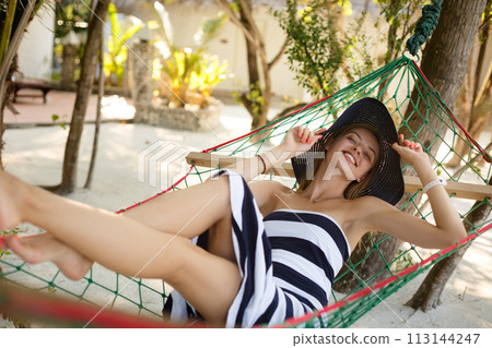 Woman relaxing in the hammock on tropical beach in the shadow, hot sunny day. Girl looks to camera with smile Woman relaxing in the hammock on tropical beach in the shadow, hot sunny day. Girl looks to camera with smile 113144247