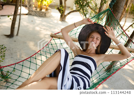 Woman relaxing in the hammock on tropical beach in the shadow, hot sunny day. Girl looks to camera with smile Woman relaxing in the hammock on tropical beach in the shadow, hot sunny day. Girl looks to camera with smile 113144248