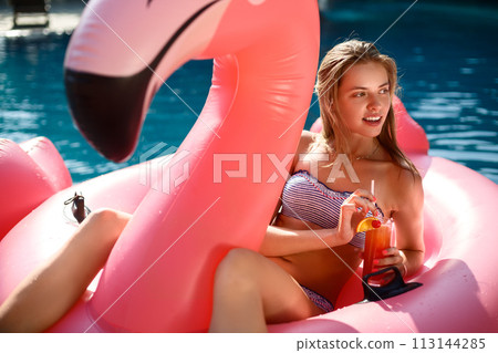 Young and sexy girl having fun and laughing on an inflatable giant pink flamingo pool float mattress with a cocktail glass. Attractive tanned woman lies in the sun on vacation 113144285