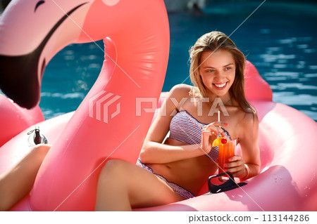 Young and sexy girl having fun and laughing on an inflatable giant pink flamingo pool float mattress with a cocktail glass. Attractive tanned woman lies in the sun on vacation Young and sexy girl having fun and laughing on an inflatable giant pink flamingo pool float mattress with a cocktail glass. Attractive tanned woman lies in the sun on vacation 113144286