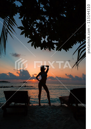 Silhouette of a beautiful slender girl on a sunset background and sun loungers on the ocean shore. Woman watch skyline on sunrise on near deck chair at the sea beach. Vacation Concept. Silhouette of a beautiful slender girl on a sunset background and sun loungers on the ocean shore. Woman watch skyline on sunrise on near deck chair at the sea beach. Vacation Concept. 113144310