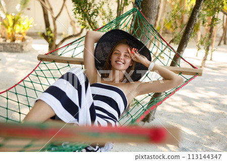 Woman relaxing in the hammock on tropical beach in the shadow, hot sunny day. Girl looks to camera with smile 113144347