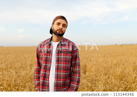 Portrait of a bearded farmer standing in a wheat field. Stilish hipster man with trucker hat and checkered shirt on. Agricultural worker Portrait of a bearded farmer standing in a wheat field. Stilish hipster man with trucker hat and checkered shirt on. Agricultural worker 113144348