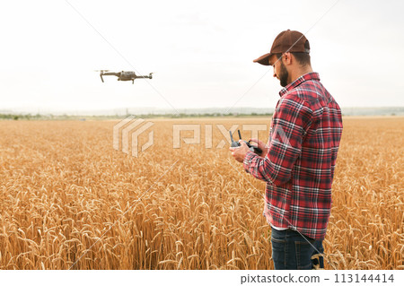 Farmer holds remote controller with his hands while quadcopter is flying on background. Drone hovers behind the agronomist in wheat field. Agricultural new technologies and innovations. Back view 113144414