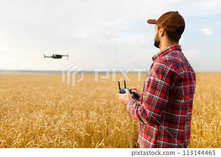 Farmer holds remote controller with his hands while quadcopter is flying on background. Drone hovers behind the agronomist in wheat field. Agricultural new technologies and innovations. Back view Farmer holds remote controller with his hands while quadcopter is flying on background. Drone hovers behind the agronomist in wheat field. Agricultural new technologies and innovations. Back view 113144465