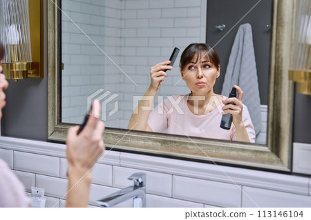 Middle-aged woman with setting spray for styling hair fixation comb looking in mirror 113146104