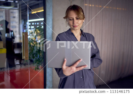 a beautiful middle age concentrated business woman wearing gray shirt and using laptop while standing in modern workspace a beautiful middle age concentrated business woman wearing gray shirt and using laptop while standing in modern workspace 113146175