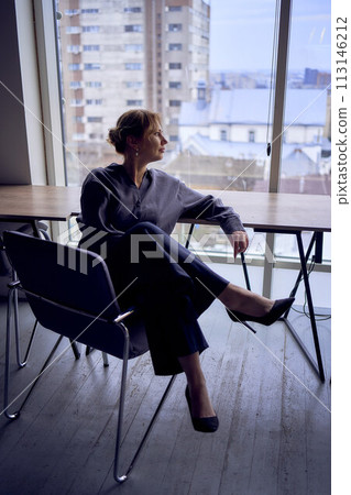 Woman sitting on chair with legs crossed over the armrest. High heels, black pants, silk blouse. Cityscape outside through window. Modern interior design with neutral tones. Woman sitting on chair with legs crossed over the armrest. High heels, black pants, silk blouse. Cityscape outside through window. Modern interior design with neutral tones. 113146212