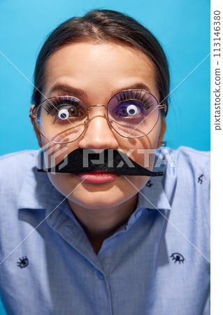 Young woman in shirt, wearing fake moustaches, glasses with eyes items on it and making funny expressions against blue background 113146380