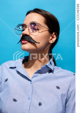 Young woman in shirt, wearing fake moustaches, glasses with eyes items on it standing with closed eyes against blue background Young woman in shirt, wearing fake moustaches, glasses with eyes items on it standing with closed eyes against blue background 113146386