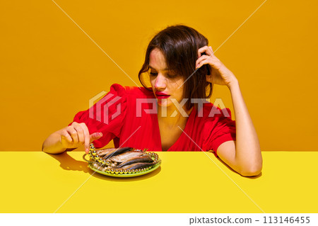 Young woman with smudged lipstick makeup eating fish against yellow background. Impact of food on health and appearance. 113146455