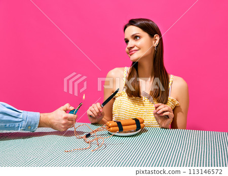 Smiling young woman sitting at table with player and sausages imitating explosive item against pink background. Surrealism Smiling young woman sitting at table with player and sausages imitating explosive item against pink background. Surrealism 113146572