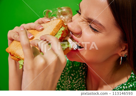Young woman eating quirky sandwich with doll against red background. Weirdness and surrealism. Close-up 113146593