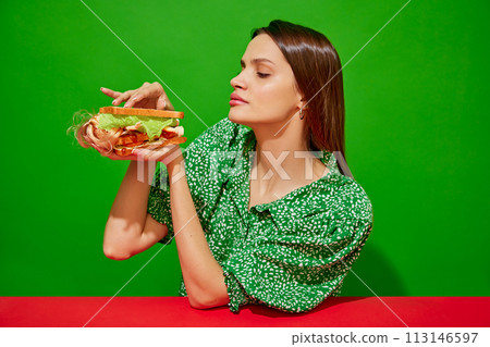 Young woman eating quirky sandwich with doll against red background. Lettuce, tomato and ketchup for additional taste Young woman eating quirky sandwich with doll against red background. Lettuce, tomato and ketchup for additional taste 113146597
