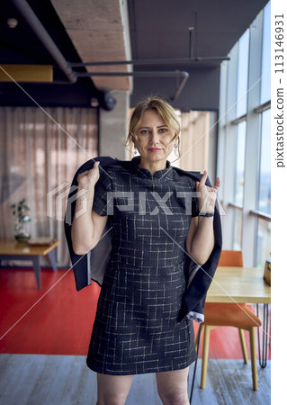 attractive woman in a black business style mini dress throws a jacket over her shoulders in a modern coworking space with panoramic windows and a black ceiling attractive woman in a black business style mini dress throws a jacket over her shoulders in a modern coworking space with panoramic windows and a black ceiling 113146931